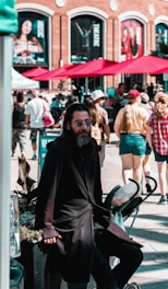 A man in dark clothing, wearing sunglasses, stands casually in an outdoor setting. There is a stroller beside him and people walking in the background, suggesting a crowded street scene. Bright red umbrellas and a brick building with large advertisements can be seen, setting a lively urban atmosphere.