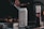 A can of Nütrl vodka soda with pineapple flavor is placed prominently in focus. It appears to have condensation, suggesting it is cold. In the background, there is a blurry image of someone grilling food, with a bottle containing sauce next to the can.