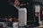 A can of Nütrl vodka soda with pineapple flavor is placed prominently in focus. It appears to have condensation, suggesting it is cold. In the background, there is a blurry image of someone grilling food, with a bottle containing sauce next to the can.