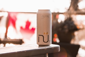A can of NÜTRL vodka soda is prominently placed on a wooden surface with a blurred Canadian flag in the background. The lighting is warm, with sunlight creating a soft, golden glow.