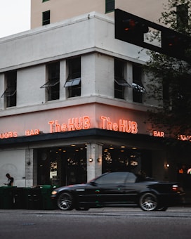 A corner building with a sign that reads The HUB in bright neon lights. A black car is driving by and a person is standing by a row of green garbage bins on the sidewalk. The building has multiple windows with awnings and is made of white brick.