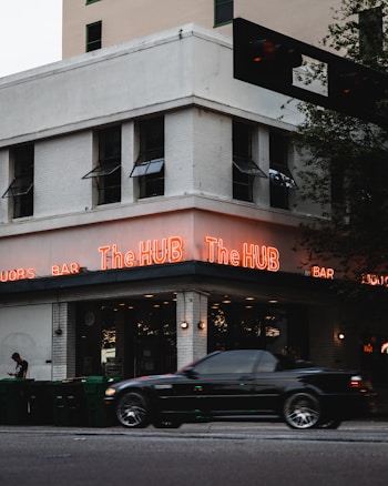 A corner building with a sign that reads The HUB in bright neon lights. A black car is driving by and a person is standing by a row of green garbage bins on the sidewalk. The building has multiple windows with awnings and is made of white brick.