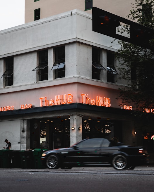 A corner building with a sign that reads The HUB in bright neon lights. A black car is driving by and a person is standing by a row of green garbage bins on the sidewalk. The building has multiple windows with awnings and is made of white brick.