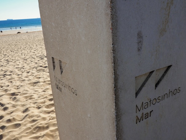 A sandy beach stretches out to meet a serene blue ocean under a clear sky. In the foreground, a concrete pillar with the inscription 'Matosinhos Mar' stands as a focal point. Distant figures can be seen walking along the shore.
