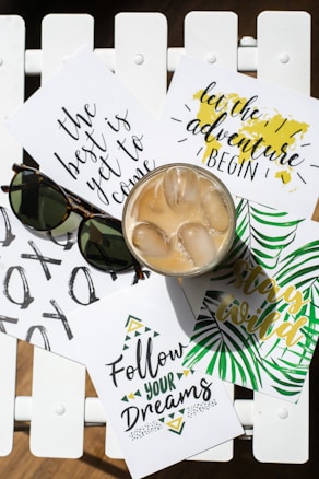 A wooden white surface holds several sheets of paper with motivational phrases like 'The best is yet to come', 'Follow your dreams', 'Stay wild', and 'Let the adventure begin'. There is a pair of sunglasses and a glass of iced coffee on top of the papers.