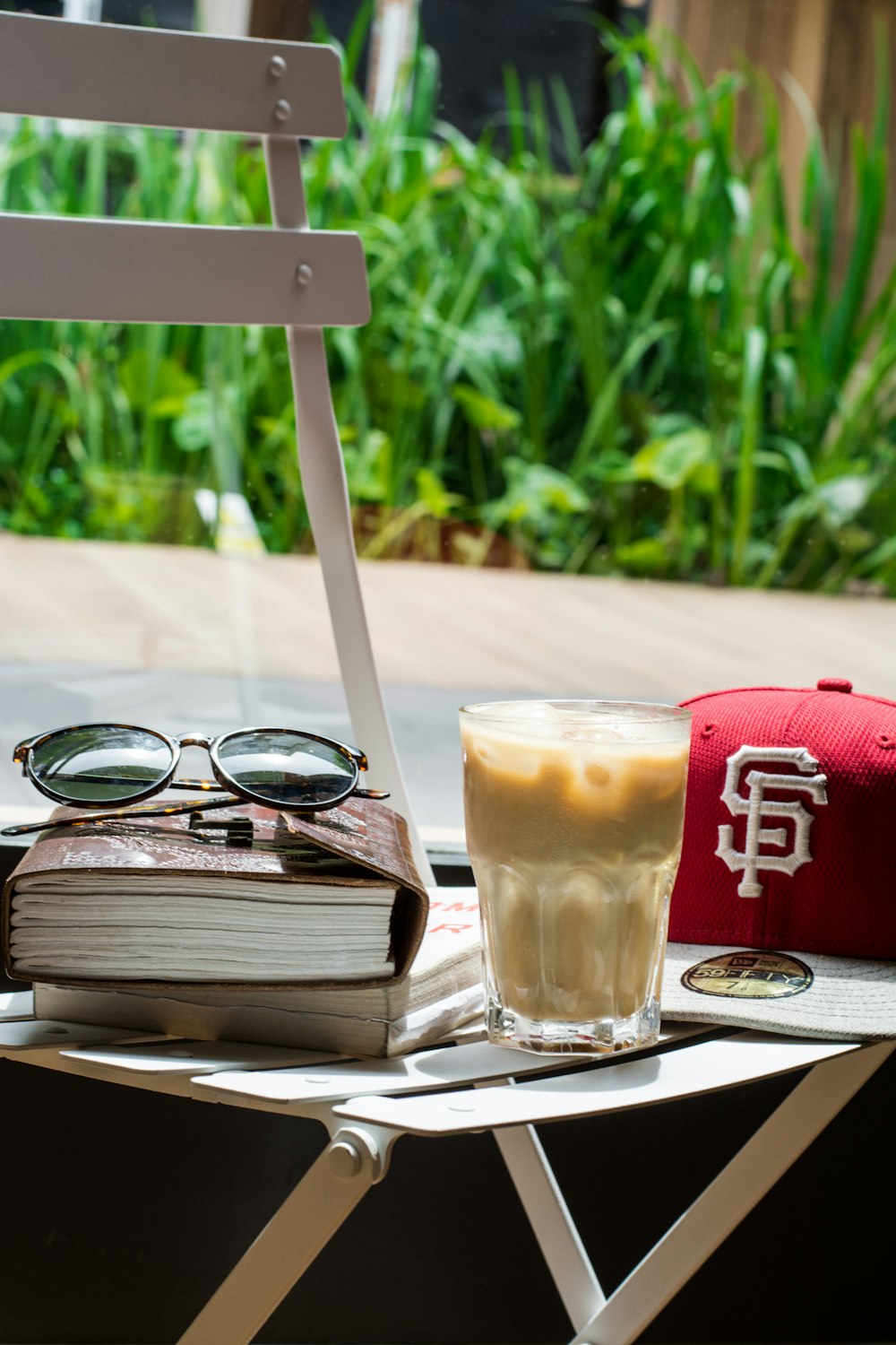 boné vermelho, copo de bebida, marrom, livro e óculos de sol pretos em cima da mesa branca