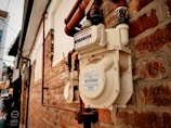 A close-up view of a gas meter attached to a red brick wall. The meter features a white exterior with visible labels and numbers. Several copper pipes are connected to the meter, extending along the wall. The background includes more brickwork and urban elements, suggesting an alley or side street.
