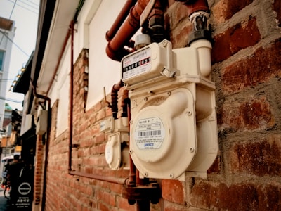 A close-up view of a gas meter attached to a red brick wall. The meter features a white exterior with visible labels and numbers. Several copper pipes are connected to the meter, extending along the wall. The background includes more brickwork and urban elements, suggesting an alley or side street.