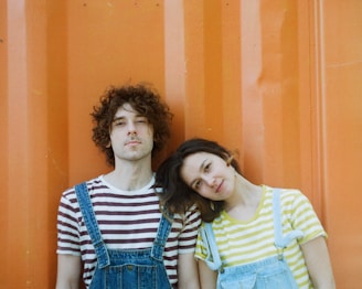A young couple stands close together against an orange background. Both wear striped shirts; one with brown and white stripes, the other with yellow and white stripes. They both wear denim overalls, and the person on the right leans their head on the other's shoulder.