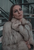 A person with subtle makeup and long, straight hair wearing a luxurious fur coat. The background consists of industrial-like structures, including metal railings and stairs, suggesting an urban setting.