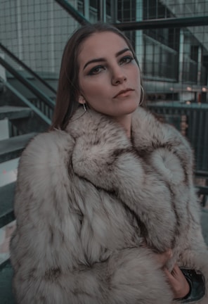 A person with subtle makeup and long, straight hair wearing a luxurious fur coat. The background consists of industrial-like structures, including metal railings and stairs, suggesting an urban setting.