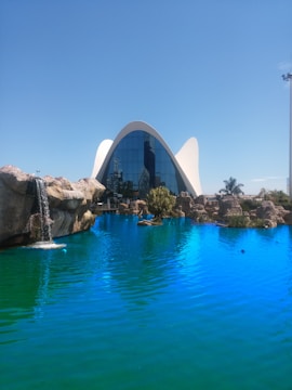A modern architectural building with a unique curved roof stands behind a vibrant blue pool of water. The building's front is made of reflective glass that mirrors the surroundings. Surrounding the pool are rocky formations and a small waterfall on the left side. The scene is tranquil and set against a clear blue sky with some greenery present.
