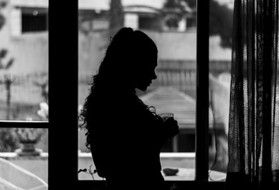 A silhouette of a person with long, curly hair is standing by the window, holding a glass. The background appears to be an outdoor scene with blurred details of a building and vegetation. Light filters through a curtain, enhancing the contrast between light and shadow.
