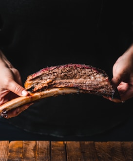 A person holds a large, grilled piece of meat with a bone, showcasing the texture and juiciness of the cut. The meat appears well-cooked with a noticeable smoky bark and is presented against a dark background, highlighting the richness of the meat.