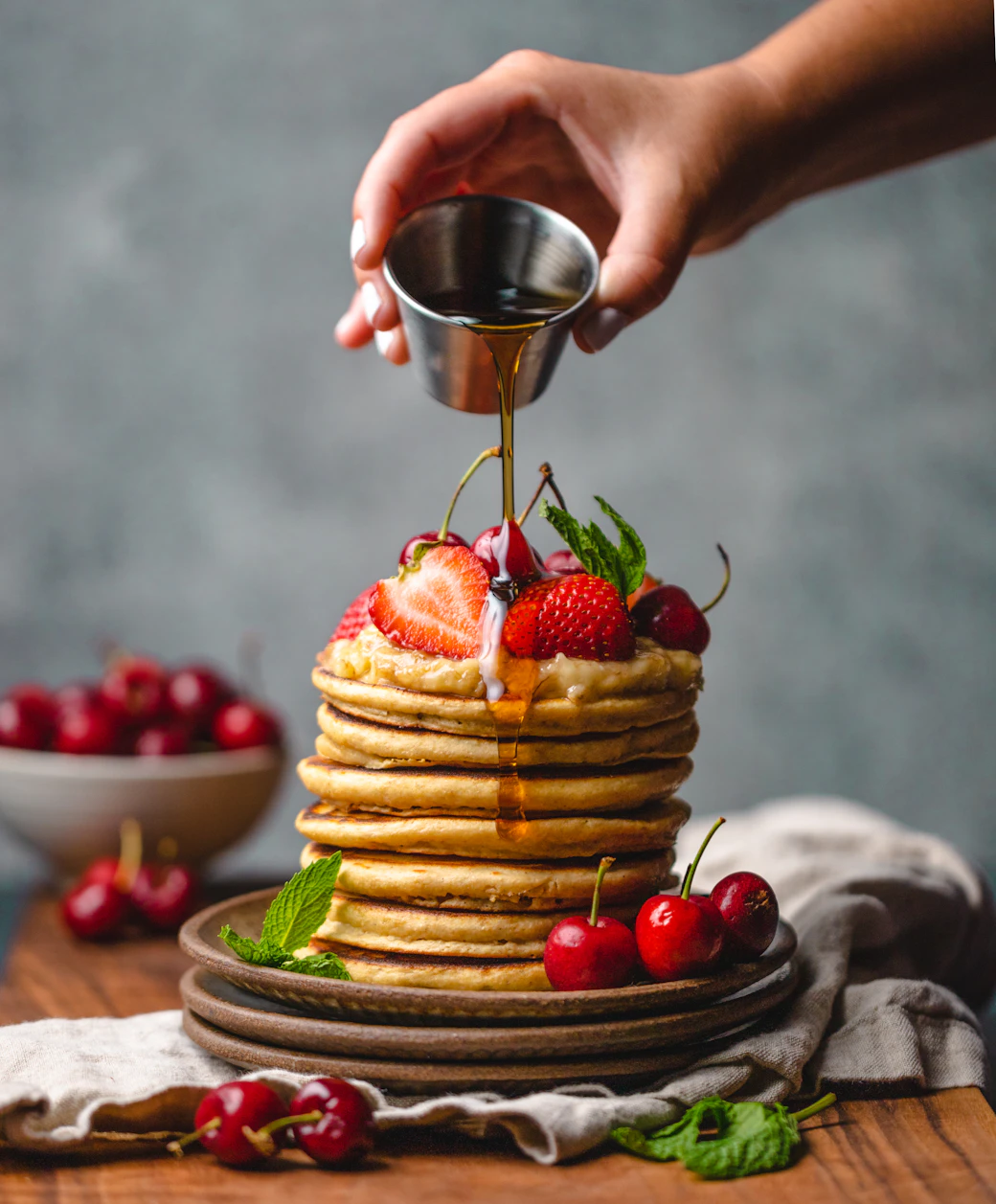 A towering stack of fluffy American pancakes crowned with strawberries and cherries, golden syrup cascading down