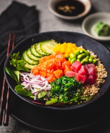 A beautifully arranged poke bowl featuring a variety of colorful ingredients including sliced cucumbers, diced orange and red fish, edamame beans, seaweed, thinly sliced onions, and brown rice. The dish is garnished with sesame seeds and accompanied by a small dish of soy sauce and a wasabi mound in the background. Chopsticks are placed adjacent to the bowl, enhancing the presentation.