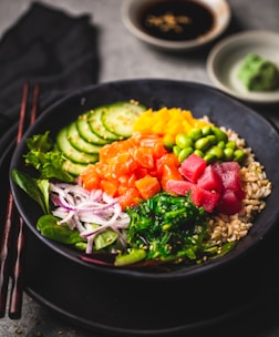 A beautifully arranged poke bowl featuring a variety of colorful ingredients including sliced cucumbers, diced orange and red fish, edamame beans, seaweed, thinly sliced onions, and brown rice. The dish is garnished with sesame seeds and accompanied by a small dish of soy sauce and a wasabi mound in the background. Chopsticks are placed adjacent to the bowl, enhancing the presentation.