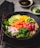 A beautifully arranged poke bowl featuring a variety of colorful ingredients including sliced cucumbers, diced orange and red fish, edamame beans, seaweed, thinly sliced onions, and brown rice. The dish is garnished with sesame seeds and accompanied by a small dish of soy sauce and a wasabi mound in the background. Chopsticks are placed adjacent to the bowl, enhancing the presentation.