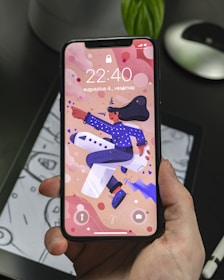 A hand holding a smartphone displaying a colorful animated wallpaper featuring a person in a blue outfit and hat riding an airplane. The background includes abstract shapes and the phone display shows the time 22:40 along with a date in another language. The phone is held over a tablet displaying a monochromatic drawing.