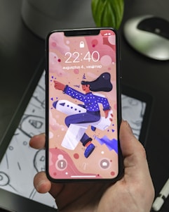 A hand holding a smartphone displaying a colorful animated wallpaper featuring a person in a blue outfit and hat riding an airplane. The background includes abstract shapes and the phone display shows the time 22:40 along with a date in another language. The phone is held over a tablet displaying a monochromatic drawing.