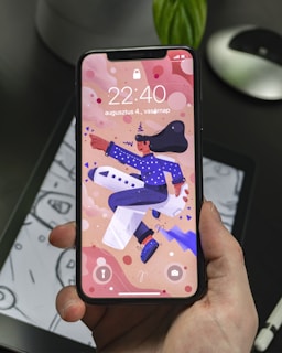 A hand holding a smartphone displaying a colorful animated wallpaper featuring a person in a blue outfit and hat riding an airplane. The background includes abstract shapes and the phone display shows the time 22:40 along with a date in another language. The phone is held over a tablet displaying a monochromatic drawing.