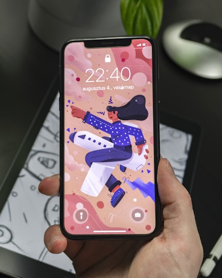 A hand holding a smartphone displaying a colorful animated wallpaper featuring a person in a blue outfit and hat riding an airplane. The background includes abstract shapes and the phone display shows the time 22:40 along with a date in another language. The phone is held over a tablet displaying a monochromatic drawing.