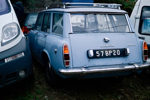 A vintage blue Simca 1500 car is parked between two other vehicles on a grassy area. The car's rear features a black license plate with the number 57BP20. The vehicle shows signs of age but remains mostly intact, with characteristic chrome details around the bumper and tail lights.