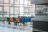 A hospital waiting area with multiple rows of brightly colored plastic chairs, including green, orange, red, and blue. The room has large windows allowing natural light to enter, and there is a reception or information desk to the right. Signage in the background indicates it's a children's hospital.