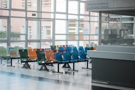 A hospital waiting area with multiple rows of brightly colored plastic chairs, including green, orange, red, and blue. The room has large windows allowing natural light to enter, and there is a reception or information desk to the right. Signage in the background indicates it's a children's hospital.