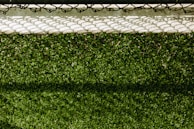 A lush green artificial turf surface is bordered at the top by a concrete edge, which has a metal chain-link fence casting a patterned shadow upon it.