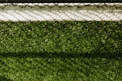 A lush green artificial turf surface is bordered at the top by a concrete edge, which has a metal chain-link fence casting a patterned shadow upon it.