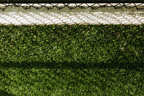 A lush green artificial turf surface is bordered at the top by a concrete edge, which has a metal chain-link fence casting a patterned shadow upon it.