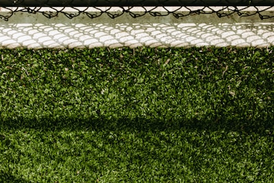 A lush green artificial turf surface is bordered at the top by a concrete edge, which has a metal chain-link fence casting a patterned shadow upon it.