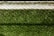 A lush green artificial turf surface is bordered at the top by a concrete edge, which has a metal chain-link fence casting a patterned shadow upon it.