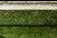 A lush green artificial turf surface is bordered at the top by a concrete edge, which has a metal chain-link fence casting a patterned shadow upon it.
