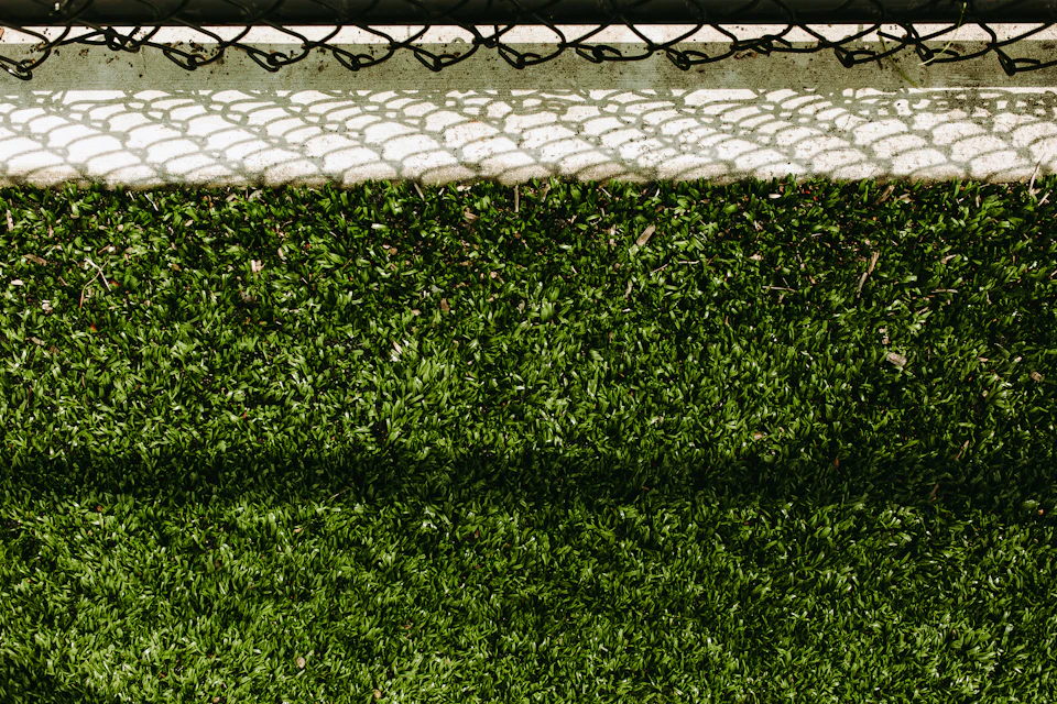 The image shows a close-up view of a tennis court, with its surface covered in lush green grass and a black chain link fence running along one side. The fence is partially visible on the right side of the frame, adding an interesting contrast to the vibrant colors of the grass. The perspective of the photo is taken from above, providing a bird's eye view of the tennis court and its surroundings.
