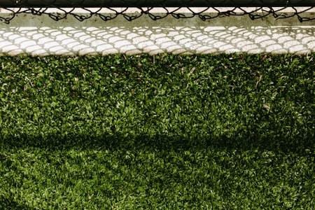 A lush green artificial turf surface is bordered at the top by a concrete edge, which has a metal chain-link fence casting a patterned shadow upon it.