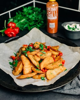 Crispy potato wedges are garnished with fresh herbs and red bell peppers, presented on parchment paper. In the background, a bottle of coconut sweet chili sauce, fresh parsley, and a small bowl of creamy dip are visible.