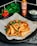 Crispy potato wedges are garnished with fresh herbs and red bell peppers, presented on parchment paper. In the background, a bottle of coconut sweet chili sauce, fresh parsley, and a small bowl of creamy dip are visible.