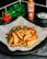 Crispy potato wedges are garnished with fresh herbs and red bell peppers, presented on parchment paper. In the background, a bottle of coconut sweet chili sauce, fresh parsley, and a small bowl of creamy dip are visible.