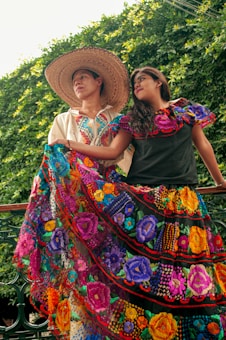 A couple dressed in traditional, colorful attire stands against a backdrop of lush greenery. The man wears a wide-brimmed straw hat and an embroidered shirt, while the woman is adorned in a bright, floral-patterned dress. They are both looking to the side with soft expressions, exuding a sense of pride and cultural richness.
