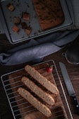 Sliced biscotti are neatly arranged on a cooling rack placed over a wooden board, with a fresh strawberry as garnish. Nearby, a serrated knife rests on the table, and a baking tray with remaining biscotti crumbs sits in the background, partially covered by a blue-gray cloth. The overall setting creates a cozy and warm baking atmosphere.