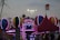 A lively amusement park scene is depicted, with brightly lit attractions featuring colorful spherical shapes and detailed patterns. A festive atmosphere is conveyed by vibrant lights, including pink and blue hues, adorning the attractions. A tall, red tent and signage suggesting a fast lane can also be seen amidst the cheerful setting, under a dimming evening sky.