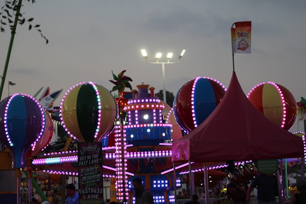 A lively amusement park scene is depicted, with brightly lit attractions featuring colorful spherical shapes and detailed patterns. A festive atmosphere is conveyed by vibrant lights, including pink and blue hues, adorning the attractions. A tall, red tent and signage suggesting a fast lane can also be seen amidst the cheerful setting, under a dimming evening sky.