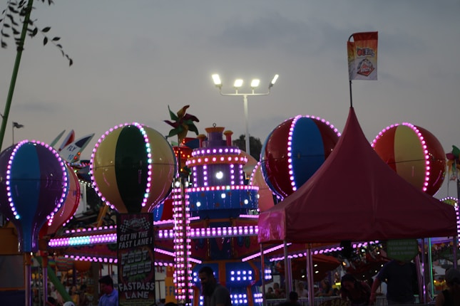 A lively amusement park scene is depicted, with brightly lit attractions featuring colorful spherical shapes and detailed patterns. A festive atmosphere is conveyed by vibrant lights, including pink and blue hues, adorning the attractions. A tall, red tent and signage suggesting a fast lane can also be seen amidst the cheerful setting, under a dimming evening sky.