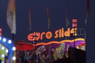 A vibrant carnival scene with a brightly lit 'Euro Slide' sign adorned with numerous lights. Below the sign, a colorful mural features cartoon-style characters with cheerful expressions. Several flags are waving in the background, adding to the festive atmosphere.