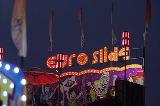 A vibrant carnival scene with a brightly lit 'Euro Slide' sign adorned with numerous lights. Below the sign, a colorful mural features cartoon-style characters with cheerful expressions. Several flags are waving in the background, adding to the festive atmosphere.