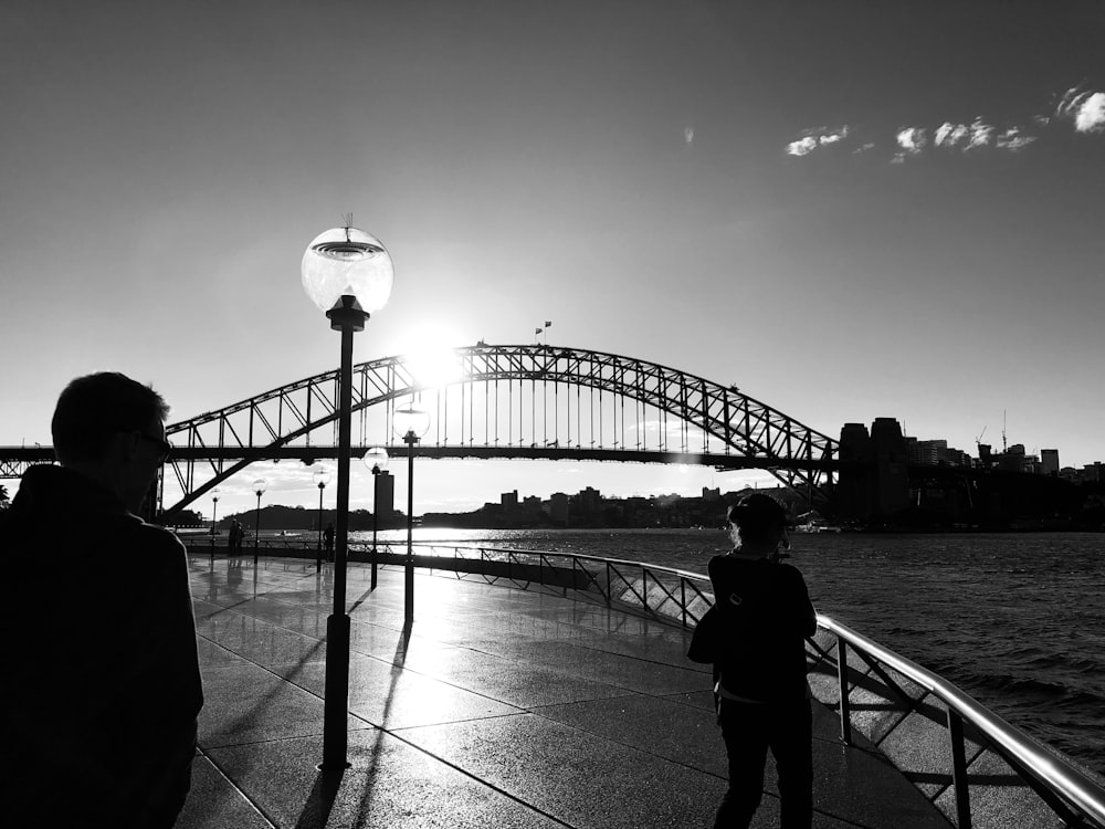 two person near bridge and body of water grey-scale photography