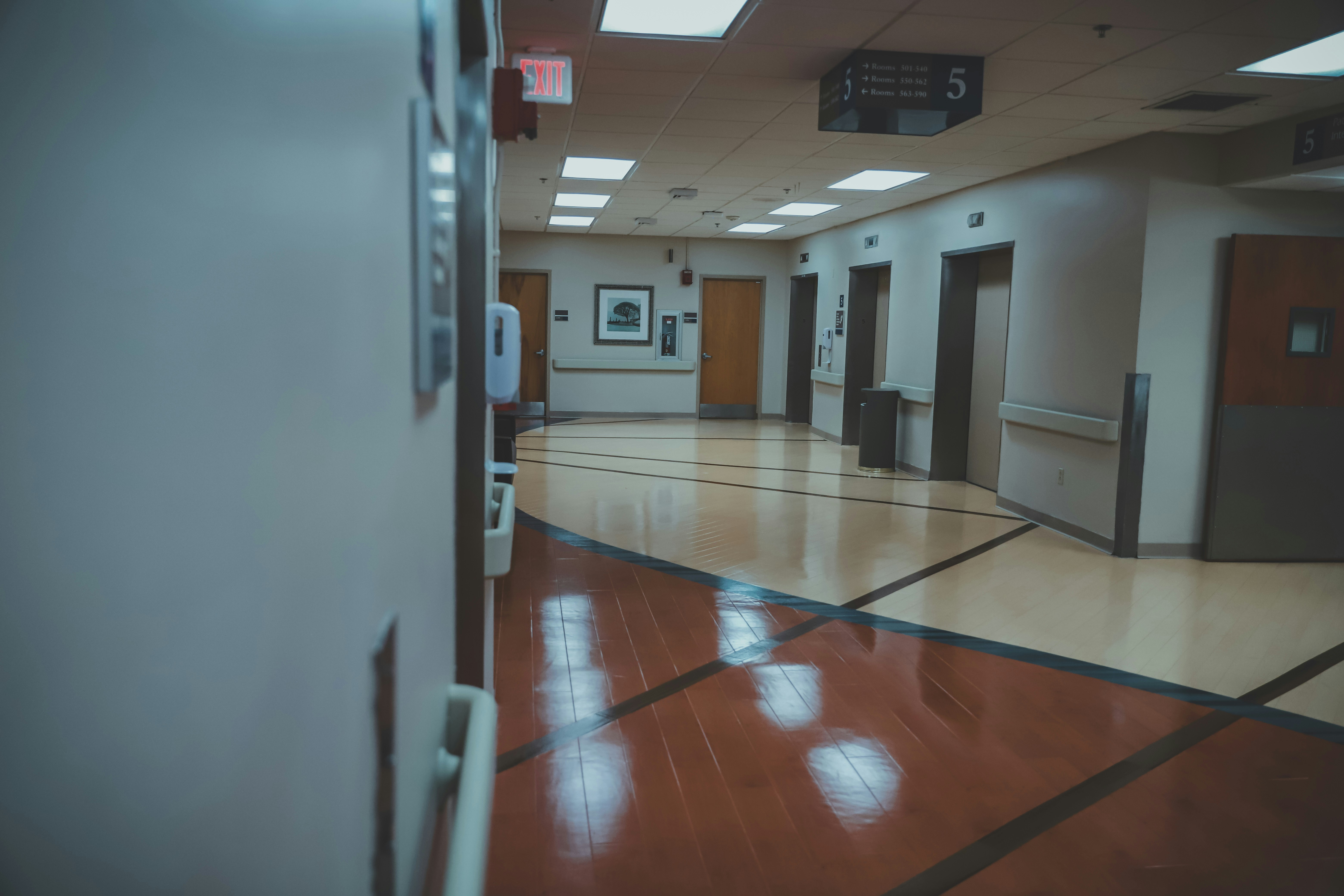 A hospital hallway with a polished floor and directional signage on the ceiling. The hallway features several doors, likely to patient rooms or offices. There is an exit sign visible and a hand sanitizer dispenser attached to the wall.