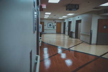 A hospital hallway with a polished floor and directional signage on the ceiling. The hallway features several doors, likely to patient rooms or offices. There is an exit sign visible and a hand sanitizer dispenser attached to the wall.